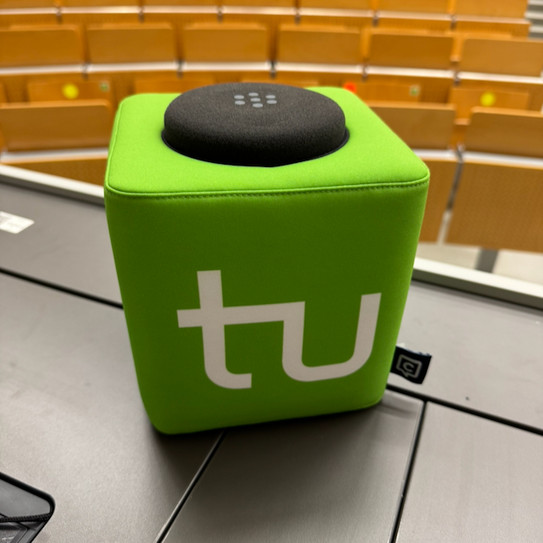 Grüner Catchbox-Mikrofonwürfel mit weißem TU-Logo auf einem Tisch in einem Hörsaal. Im Hintergrund sind die ansteigenden Sitzreihen des Hörsaals zu sehen.