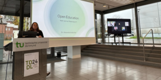 Die Keynote-Speakerin steht an einem Rednerpult mit einem Schild der Technischen Universität Dortmund (TU Dortmund) und hält einen Vortrag mit dem Titel ‘Open Education - Mehr als nur Materialien?’. Im Hintergrund ist ein großer Bildschirm zu sehen, auf dem mehrere Personen in einem Online-Meeting dargestellt sind. Der Veranstaltungsort hat große Fenster, die Tageslicht hereinlassen.