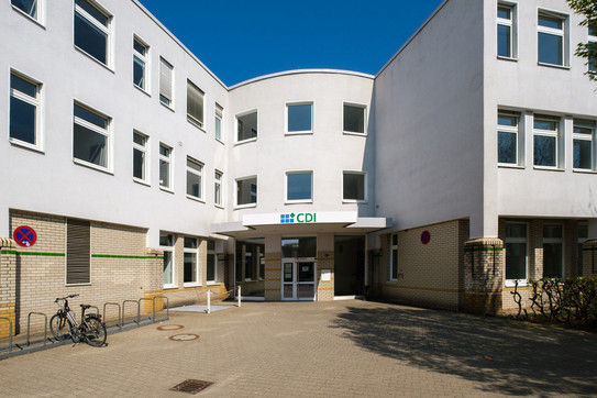 Das Foto zeigt die Fassade eines modernen Bürogebäudes mit weißen Außenwänden und großen Fenstern. Über dem Eingang befindet sich ein Schild mit dem Logo und Schriftzug ‘CDI’. Vor dem Gebäude sind Fahrradständer, ein Fahrrad sowie Verkehrsschilder zu sehen. Der Himmel ist klar und blau, was auf sonniges Wetter hinweist.